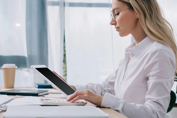 Seitenansicht einer fokussierten Geschäftsfrau mit Tablet, die am Arbeitsplatz mit Papieren im Büro Berechnungen am Taschenrechner vornimmt — Stockfoto