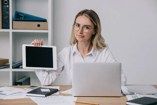 Portrait of businesswoman in eyeglasses showing tablet with blank screen at workplace with papers and laptop in office — Stock Photo