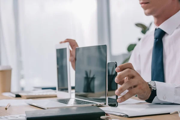 Recortado tiro de hombre de negocios mostrando portátil, tableta y teléfono inteligente con pantallas en blanco en el lugar de trabajo en la oficina - foto de stock