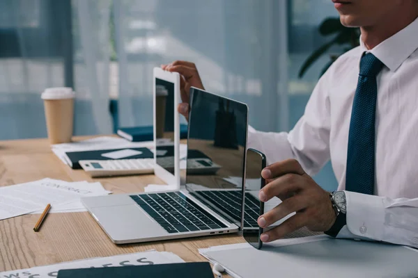 Recortado tiro de hombre de negocios mostrando portátil, tableta y teléfono inteligente con pantallas en blanco en el lugar de trabajo en la oficina - foto de stock