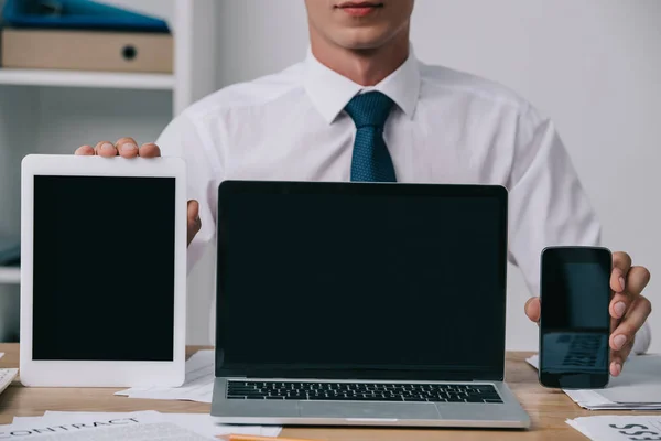 Recortado tiro de hombre de negocios mostrando portátil, tableta y teléfono inteligente con pantallas en blanco en el lugar de trabajo en la oficina — Stock Photo