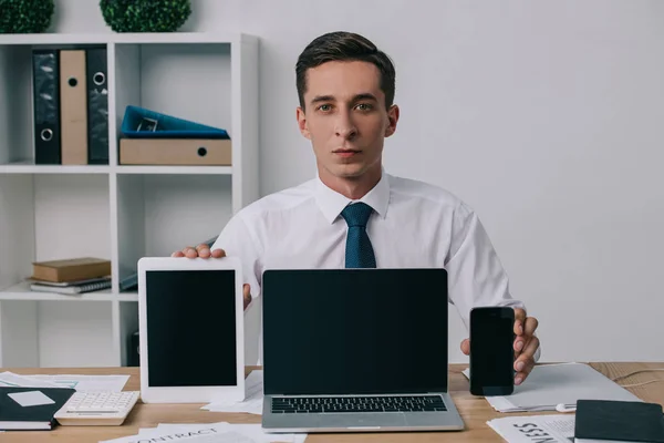 Portrait d'un homme d'affaires montrant un ordinateur portable, une tablette et un smartphone avec des écrans vides sur le lieu de travail au bureau — Photo de stock