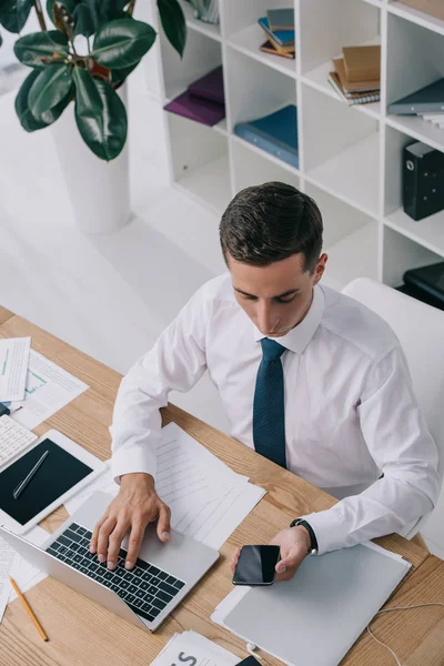Vista de ángulo alto del hombre de negocios utilizando teléfono inteligente y portátil en el lugar de trabajo con documentos en la oficina - foto de stock
