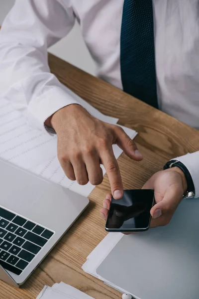 Partial view of businessman using smartphone at tabletop with documents and laptop — Stock Photo