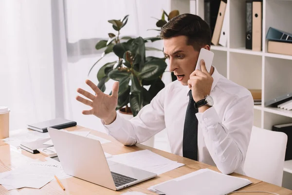 Эмоциональный бизнесмен жестом во время разговора на смартфоне на рабочем месте в офисе — стоковое фото