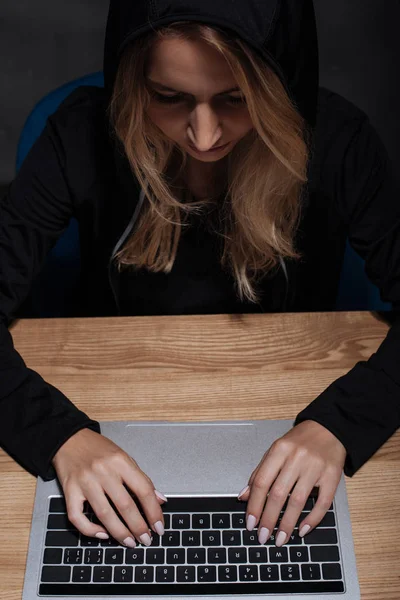 Hacker femenino en sudadera con capucha negra usando el ordenador portátil en la mesa de madera - foto de stock