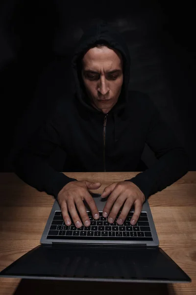 Focused hacker in black hoodie using laptop at wooden tabletop, cuber security concept — Stock Photo