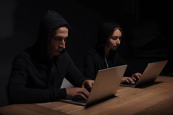 Hackers enfocados en sudaderas con capucha negra usando computadoras portátiles en habitación oscura, concepto de seguridad cibernética - foto de stock