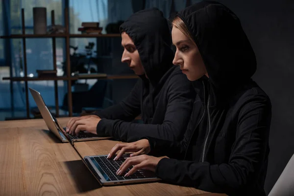 Vista lateral de los piratas informáticos en sudaderas con capucha negro utilizando computadoras portátiles en la mesa de madera, concepto de seguridad cibernética — Stock Photo