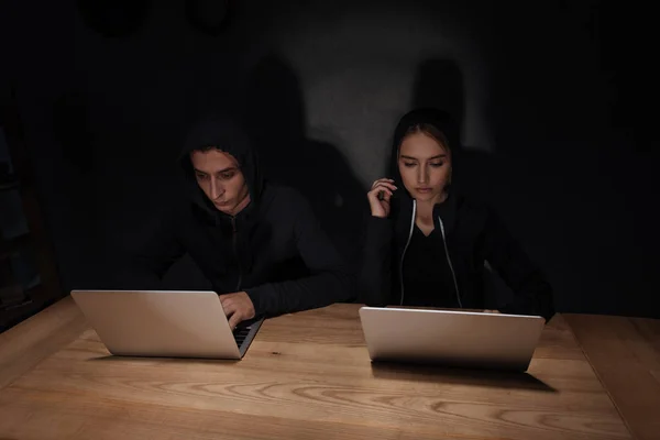 Hackers in black hoodies using laptops at wooden tabletop in dark room, cyber security concept — Stock Photo