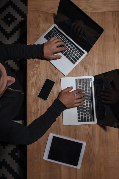 Vista aérea de hacker en sudadera con capucha negra usando computadoras portátiles en la mesa con teléfono inteligente y tableta, concepto de seguridad cibernética - foto de stock