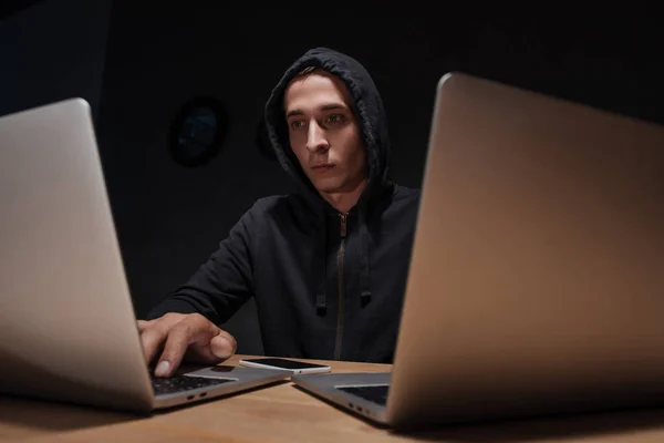 Portrait of hacker in black hoodie using laptops at tabletop with smartphone, cyber security concept — Stock Photo