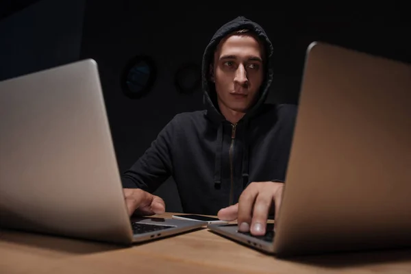 Ritratto di hacker in felpa nera con cappuccio utilizzando computer portatili in camera oscura, concetto di sicurezza informatica — Foto stock