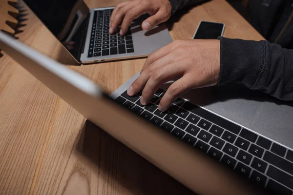 Частковий вид хакера з використанням ноутбуків на стільниці зі смартфоном, концепція кібербезпеки — стокове фото
