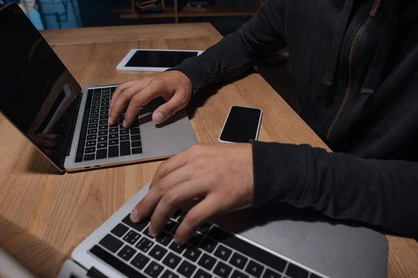 Vista parcial del hacker masculino usando computadoras portátiles en la mesa con teléfono inteligente y tableta, concepto de seguridad cibernética - foto de stock