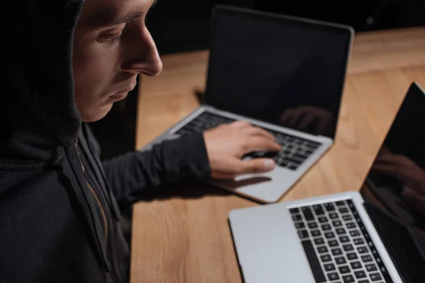 Vista lateral del hacker masculino en sudadera con capucha negra usando computadoras portátiles, concepto de seguridad cibernética - foto de stock