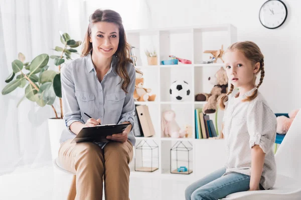 Smiling psychologist with clipboard sitting in office with little child and looking at camera — Stock Photo