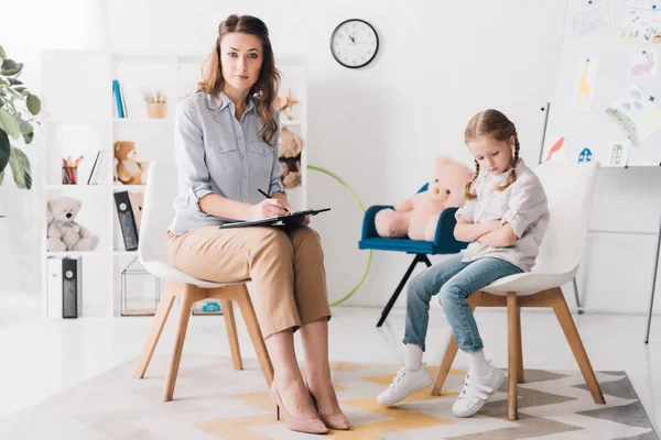 Psychologe mit Klemmbrett sitzt mit kleinem Kind im Büro und blickt in die Kamera — Stockfoto