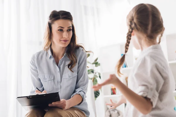 Belle psychologue adulte avec presse-papiers parlant à un petit enfant au bureau — Photo de stock