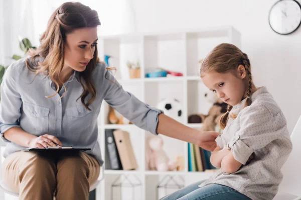 Psicólogo adulto con portapapeles hablando con un niño triste en la oficina - foto de stock