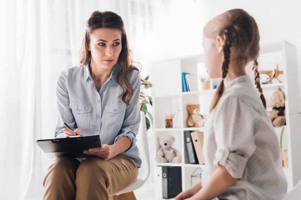 Adult psychologist with clipboard talking to little child in office — Stock Photo