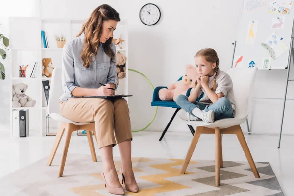 Ernsthafter Erwachsenenpsychologe mit Klemmbrett sitzt neben kleinem depressiven Kind im Büro — Stockfoto