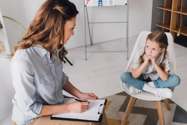 Psicologo adulto con appunti seduto vicino a un bambino depresso in ufficio — Foto stock