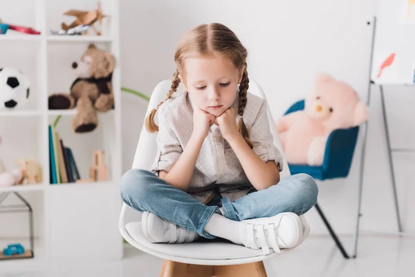 Lonely little child sitting on chair in front of shelves with toys and looking down — Stock Photo
