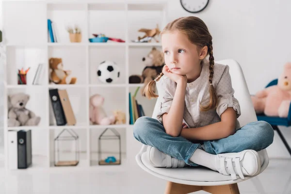 Petit enfant solitaire assis sur une chaise devant des étagères avec des jouets et regardant loin — Photo de stock