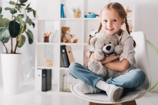 Happy little child sitting on chair and embracing her teddy bear while looking at camera — Stock Photo
