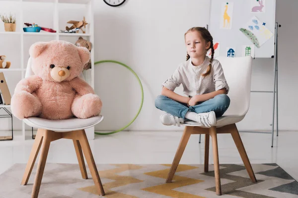 Lonely little child sitting on chair near pink teddy bear — Stock Photo