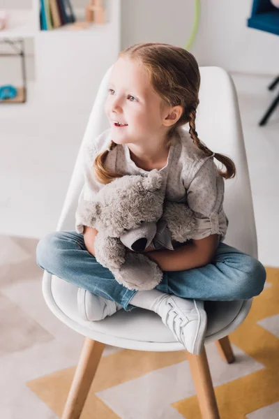 Niño pequeño y feliz sentado en la silla y abrazando su osito de peluche - foto de stock