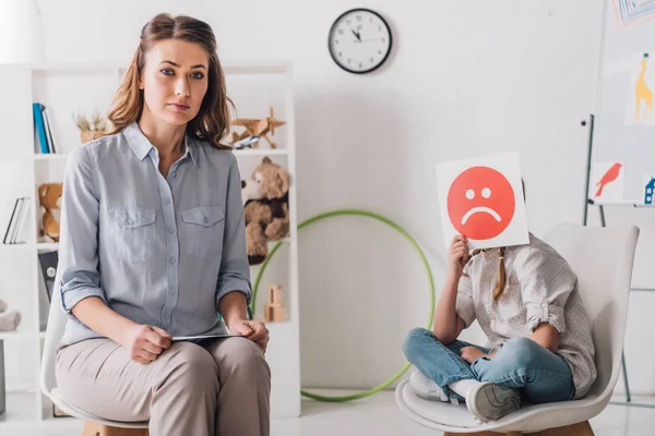 Psychologue assis près de petit enfant alors qu'elle couvre le visage avec la carte visage émotion triste — Photo de stock