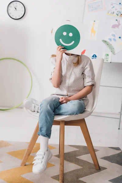 Petit enfant assis sur une chaise et couvrant le visage avec un visage souriant symbole — Photo de stock