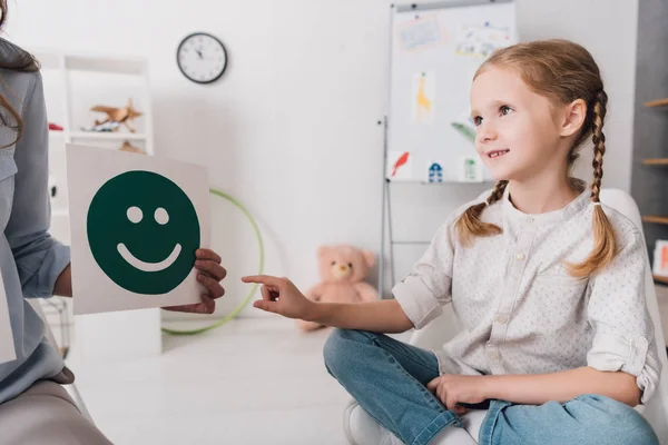 Adorable little child pointing at smiley face on paper in hand of psychologist — Stock Photo