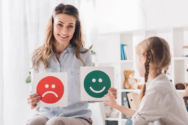 Smiling psychologist showing happy and sad emotion faces cards to child — Stock Photo