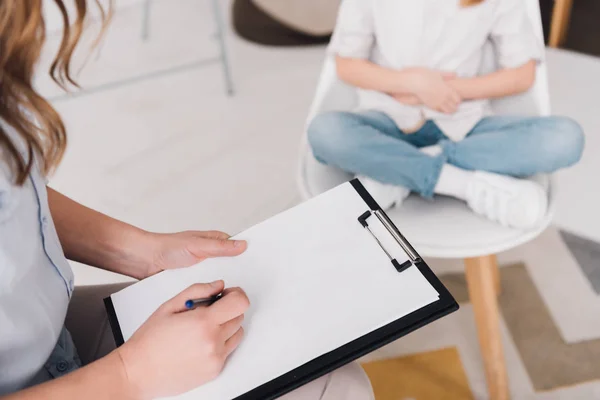 Cropped shot of psychologist with clipboard sitting in front of little child in office — Stock Photo