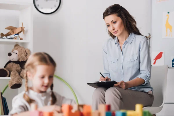 Little child playing with blocks while psychologist sitting blurred on background — Stock Photo