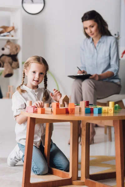 Criança pequena feliz brincando com blocos e olhando para a câmera enquanto psicólogo sentado embaçado no fundo — Fotografia de Stock