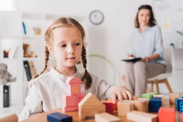 Adorable niño pequeño jugando con bloques mientras psicólogo sentado borroso en el fondo - foto de stock