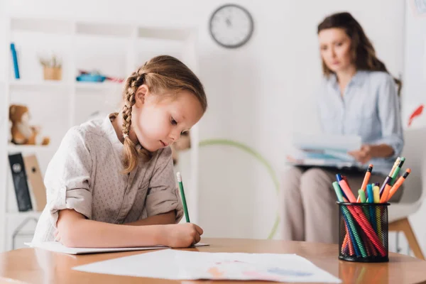 Sérieux petit enfant dessin tandis que psychologue assis flou sur le fond — Photo de stock