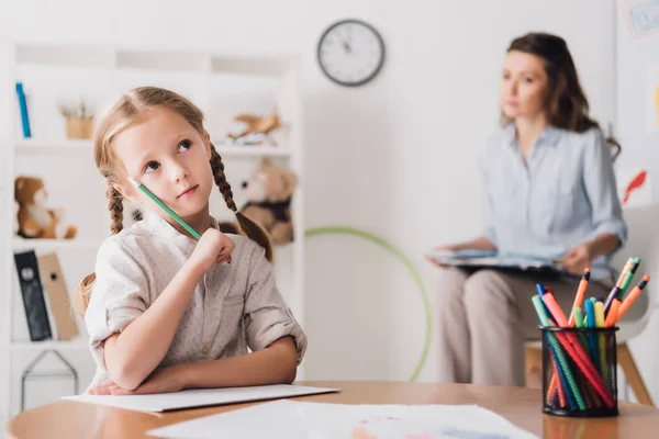 Pensoso bambino disegno mentre psicologo seduta offuscata su sfondo — Foto stock