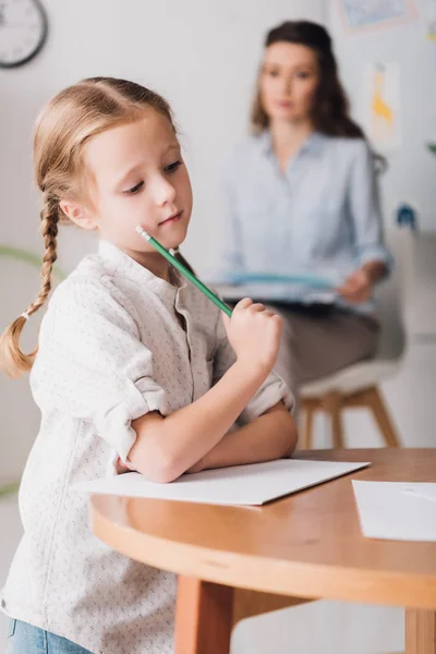 Nachdenkliches kleines Kind zeichnet, während Psychologe mit Klemmbrett unscharf auf Hintergrund sitzt — Stockfoto