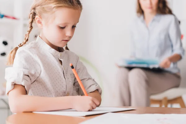 Konzentriertes kleines Kind zeichnet, während Psychologe mit Klemmbrett unscharf auf Hintergrund sitzt — Stockfoto