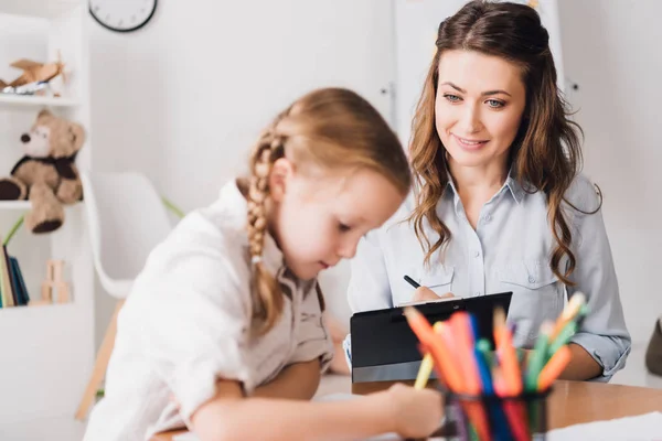 Adult psychologist writing in clipboard while looking at little drawing child — Stock Photo