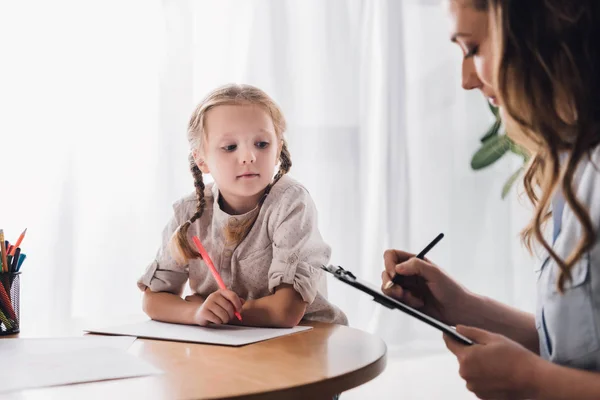 Psicólogo escribiendo en portapapeles mientras está sentado cerca de niño pequeño dibujo - foto de stock