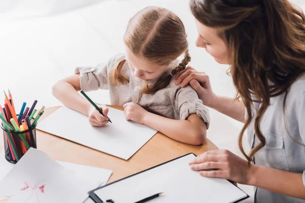 Vue grand angle de psychologue avec presse-papiers assis près du petit enfant alors qu'elle dessine avec des crayons de couleur — Photo de stock