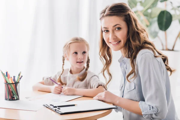 Psicologa sorridente con appunti seduta vicino al bambino mentre disegna con matite a colori e guarda la macchina fotografica — Stock Photo