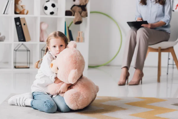 Criança assustada com ursinho sentado no chão com psicólogo sentado no fundo — Fotografia de Stock