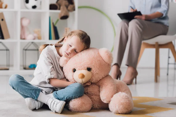 Petit enfant solitaire avec ours en peluche assis sur le sol avec psychologue assis sur le fond — Photo de stock